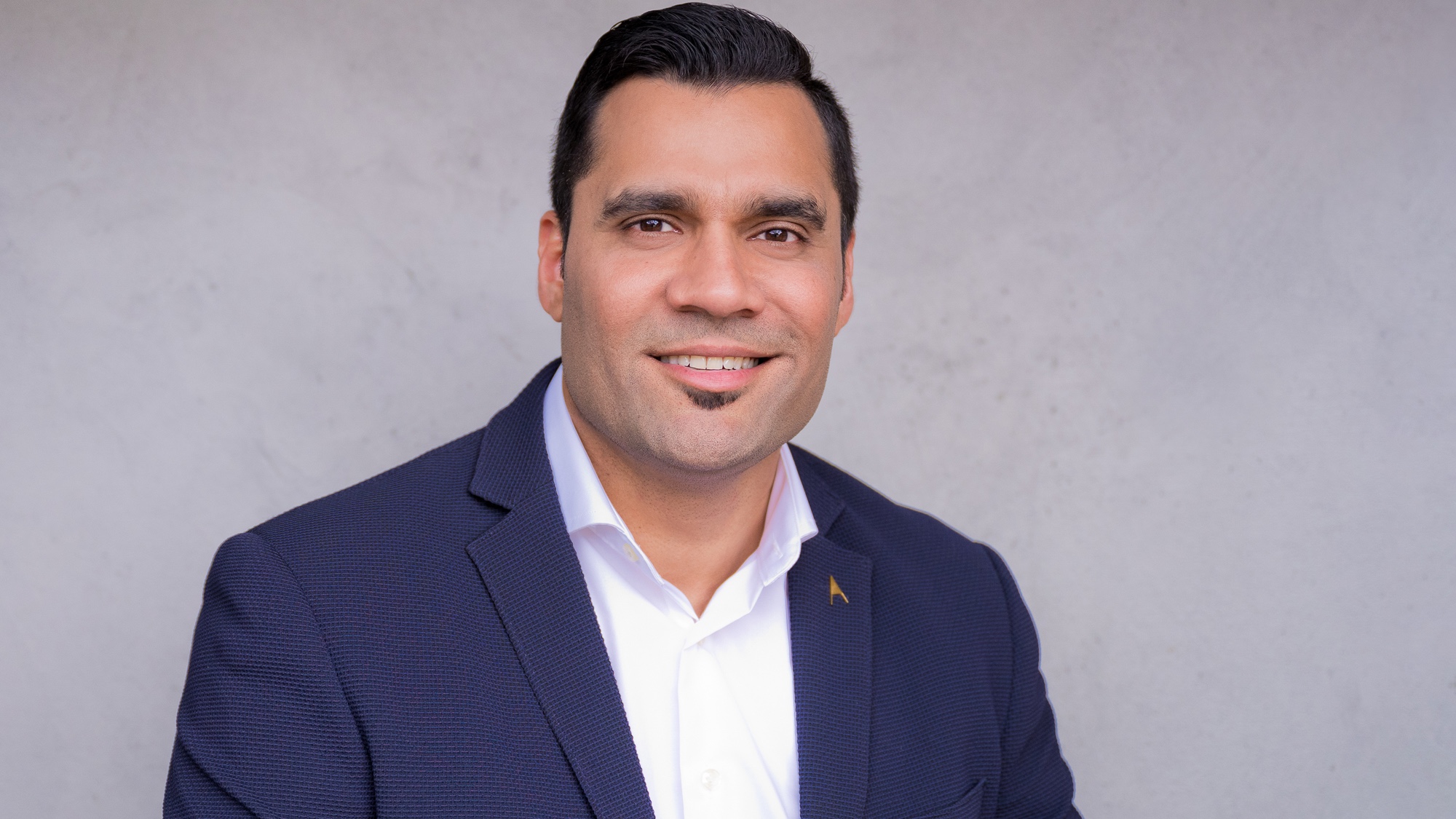 A man in a navy blue blazer and white shirt is smiling against a plain, light-colored background.