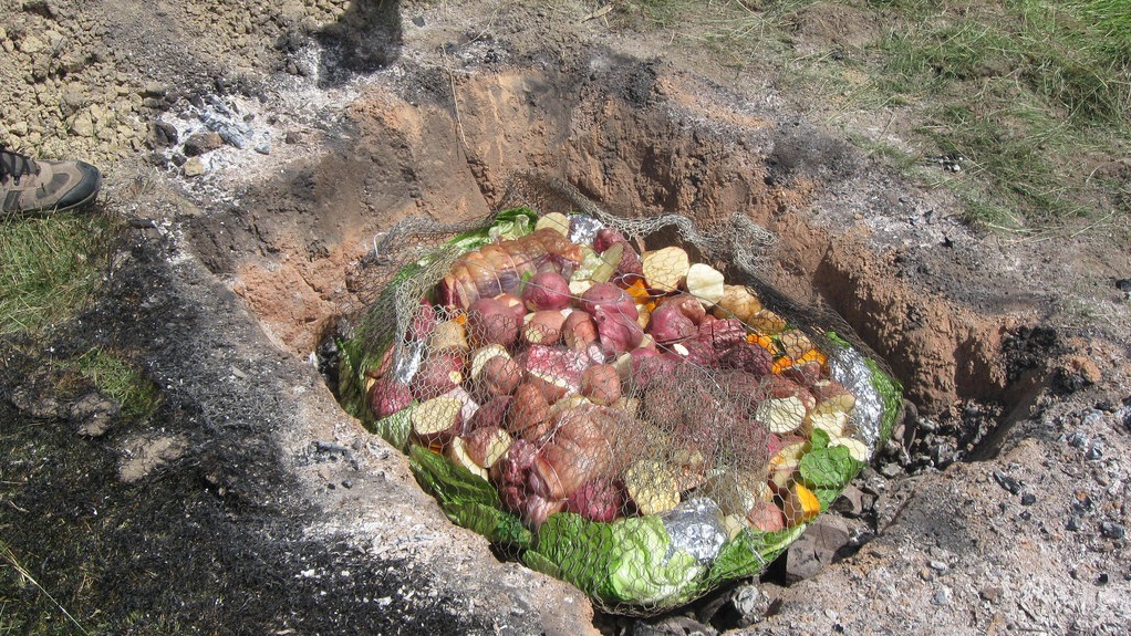 A pit filled with various vegetables, including potatoes, onions, and leafy greens, wrapped in a net and placed on hot coals for cooking.