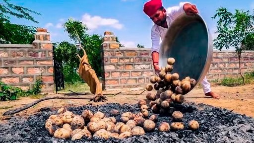 A man in a red turban and white outfit pours a large quantity of potatoes from a metal bowl onto a bed of coals, with a brick wall and greenery in the background.
