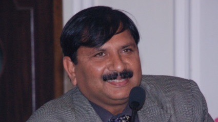 A man with a mustache is sitting in front of a microphone, wearing a gray suit and a patterned tie.