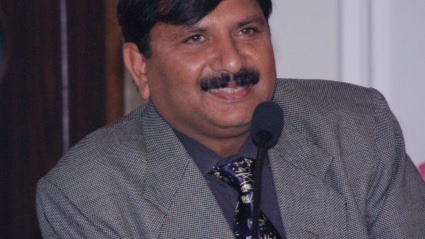 A man with a mustache is sitting in front of a microphone, wearing a gray suit and a patterned tie.