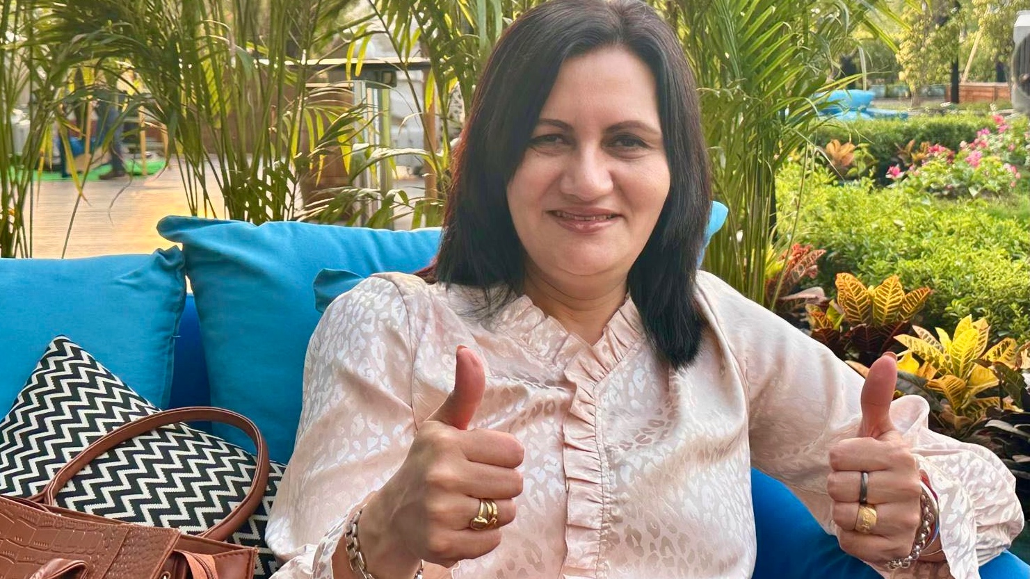 A woman sits on a blue couch, giving a thumbs up with both hands, surrounded by lush greenery and a brown handbag.
