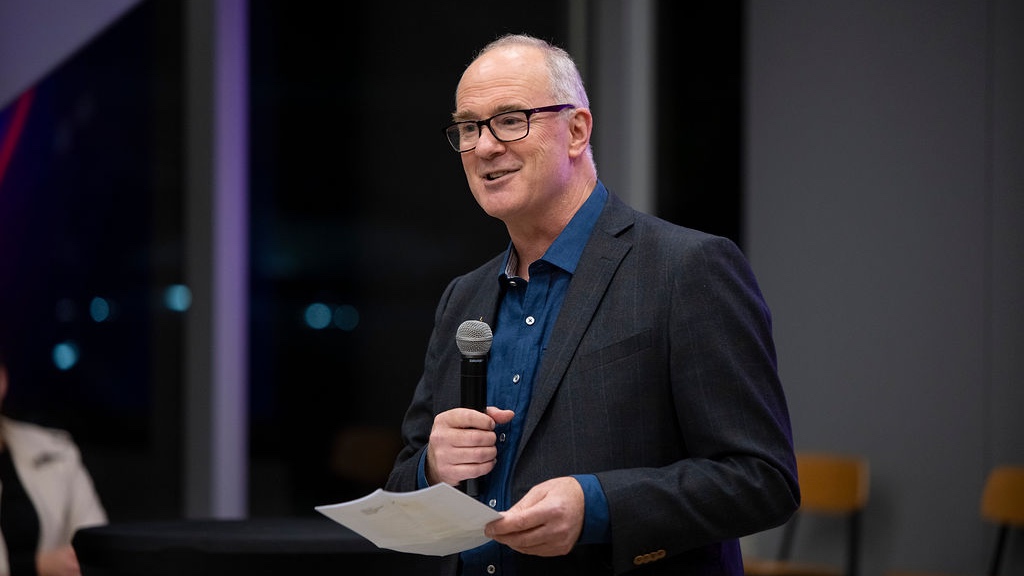 A man in a suit and glasses is holding a microphone and a piece of paper, smiling and speaking at an event.