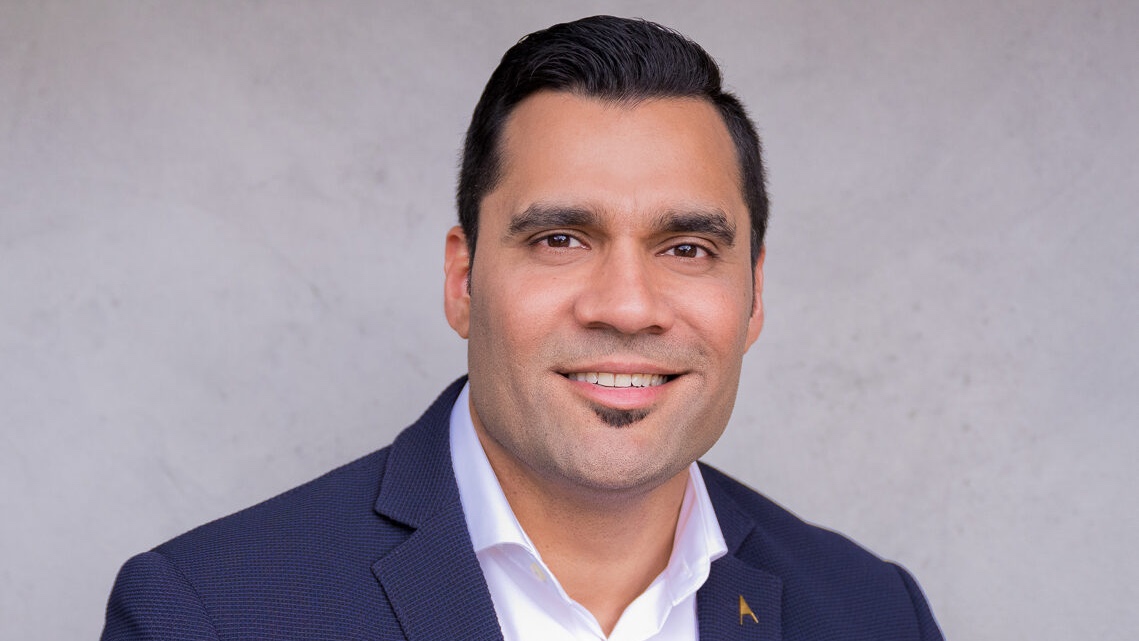 A man with a mustache and beard is smiling and wearing a navy blue suit jacket over a white shirt.