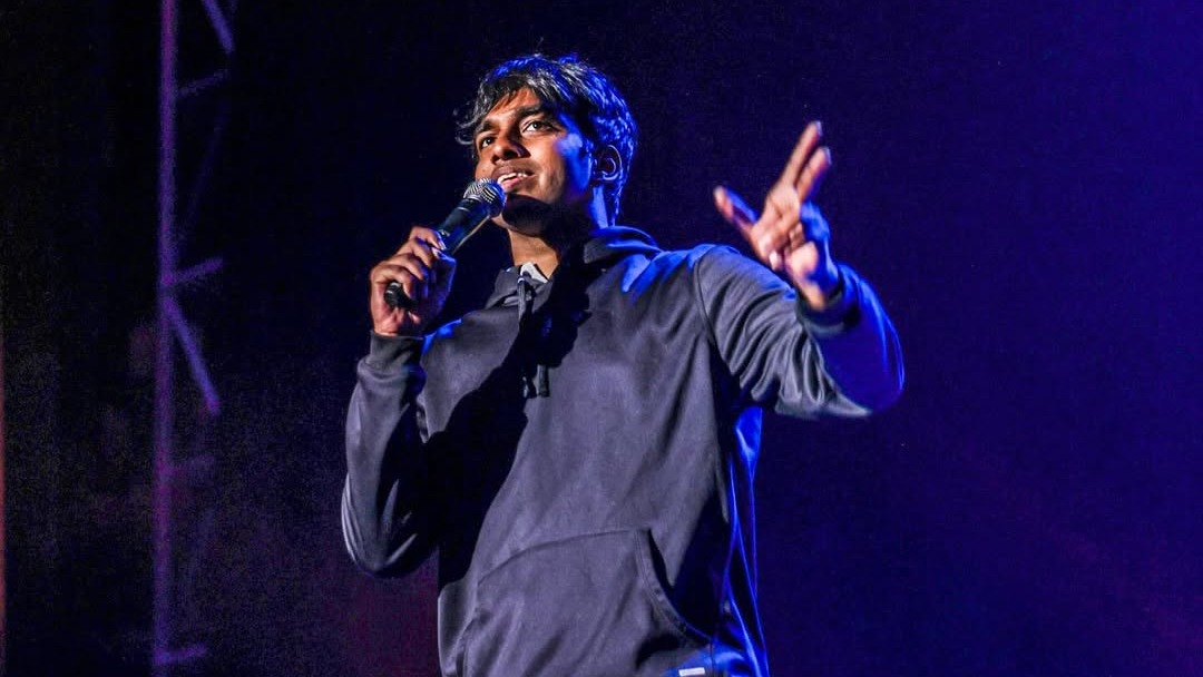 A man in a black hoodie and blue jeans is standing on stage, holding a microphone in his left hand and gesturing with his right hand.
