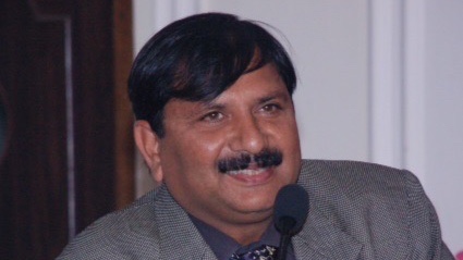 A man with a mustache and wearing a gray suit and tie is smiling and speaking into a microphone.