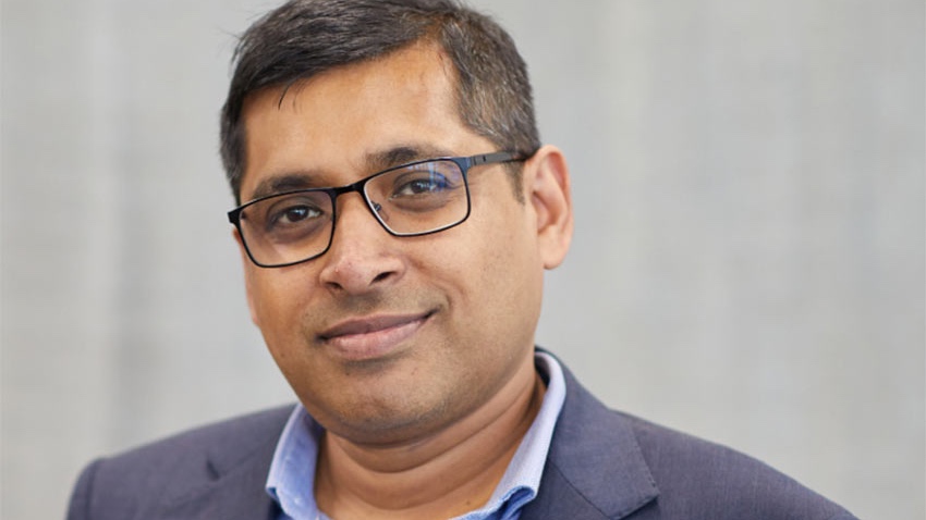 A man with short hair and glasses, wearing a blue shirt and gray blazer, smiles at the camera against a plain background.