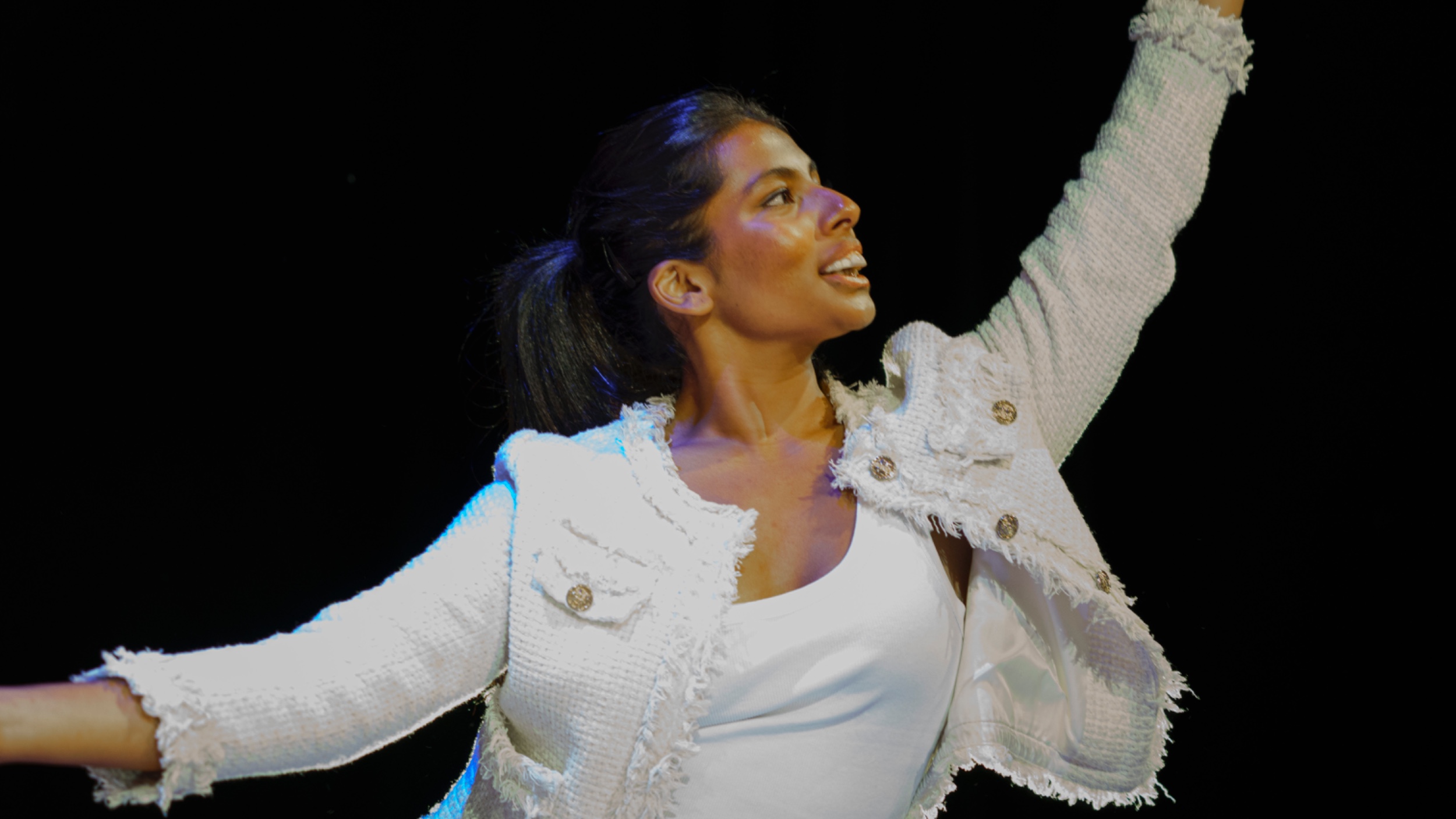 A woman in a white outfit is performing on stage, holding a cluster of blue balloons in one hand while her other hand is extended outward.