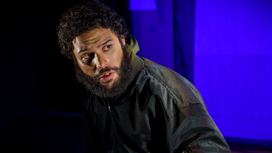 A man with a beard and curly hair, wearing a black jacket, stands in a dimly lit room with a blue curtain in the background.