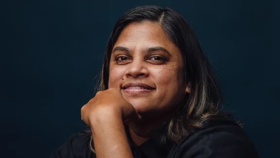 A woman with long hair and a black shirt is smiling and posing for a photo with her hand on her chin.
