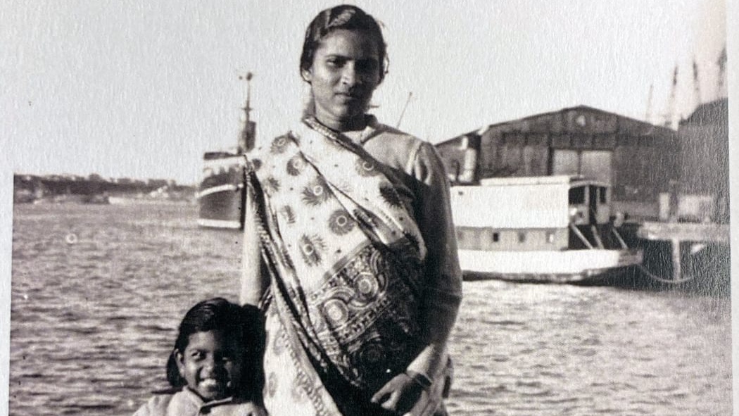 A black and white photograph of a woman and a child standing by the water, with a boat and a building in the background.