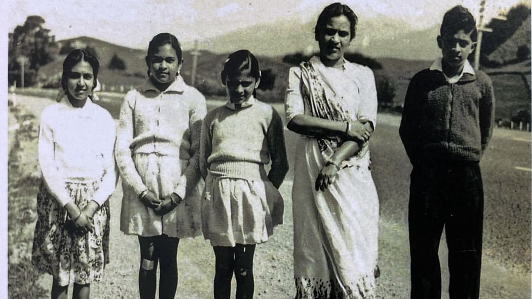 A black and white photograph of four young girls, a woman, and a boy standing on a road, with a hill and trees in the background.