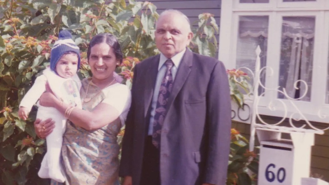 An elderly man and woman stand outside a house with a young child, with the number 60 visible on a nearby post.