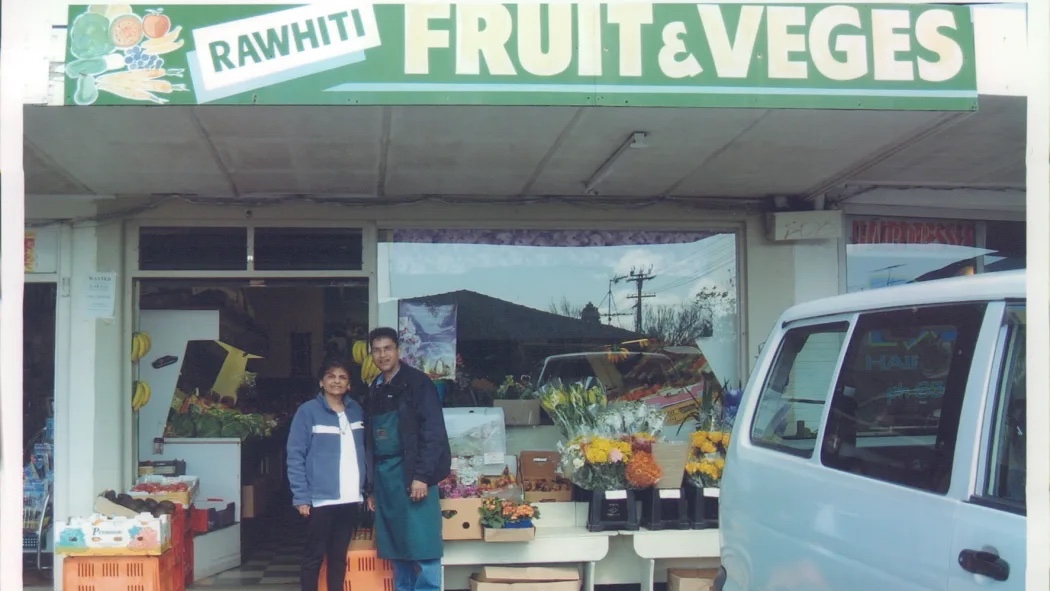 Two individuals stand outside a fruit and vegetable shop, with a white van parked nearby and various produce displayed in crates and on tables.