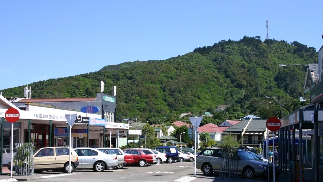 A parking lot with several cars parked in front of a row of shops, with a large green hill in the background.
