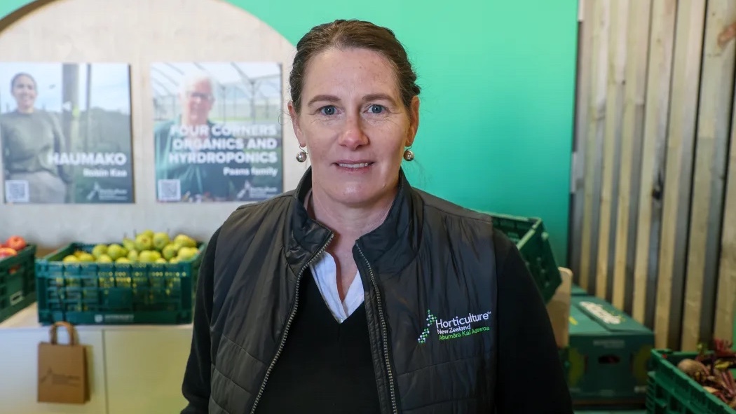 A woman stands in front of a green wall with posters, wearing a black vest with