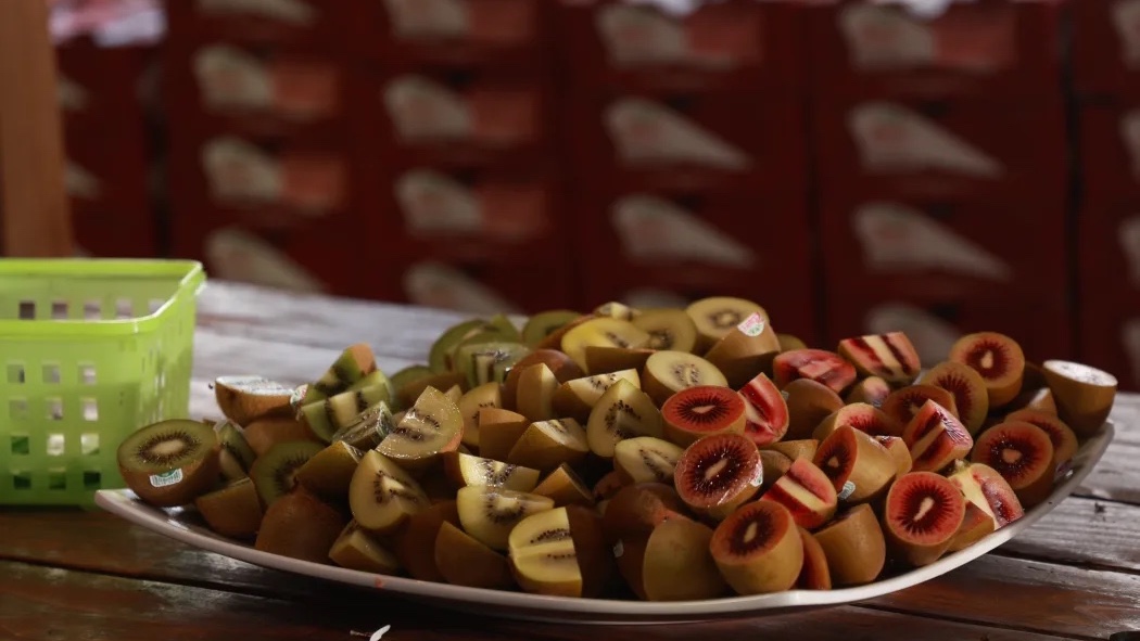 A plate filled with sliced kiwis and apples is placed on a wooden table next to a green basket.