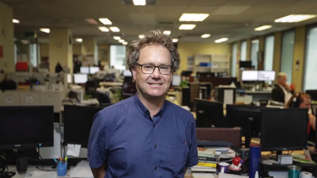 A man with glasses and a blue shirt stands in a busy office, surrounded by multiple desks with computers, papers, and office supplies.