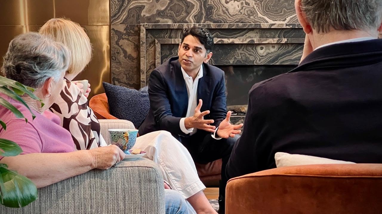 A man in a suit is speaking to a group of people seated in a living room, with a fireplace in the background.