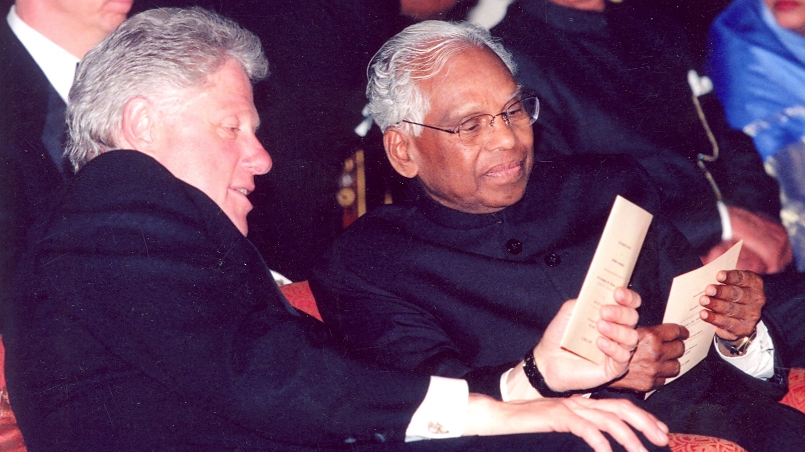 Two men, one wearing glasses, are seated on a red couch, holding papers and smiling, while several other individuals are seated behind them.