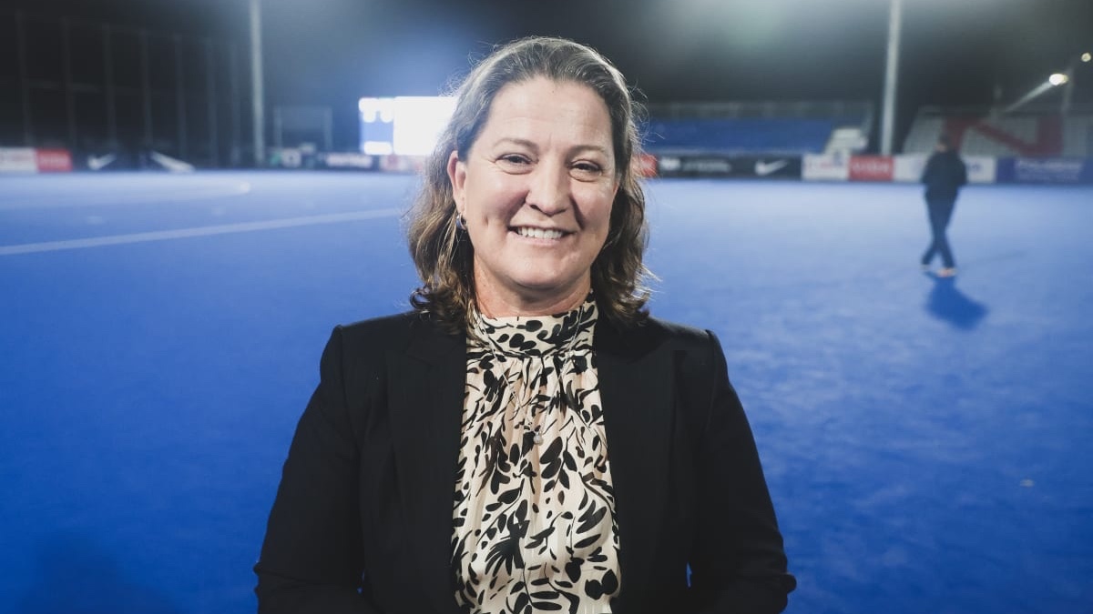 A woman in a black blazer and patterned blouse stands on a blue sports field at night, smiling at the camera with a person walking in the background.
