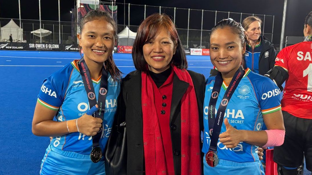 Two female athletes pose with a woman, all smiling and holding medals, on a blue sports field with other team members in the background.