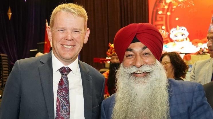 Two men, one in a suit and tie and the other in a blue suit with a red turban, stand smiling next to each other at an event.